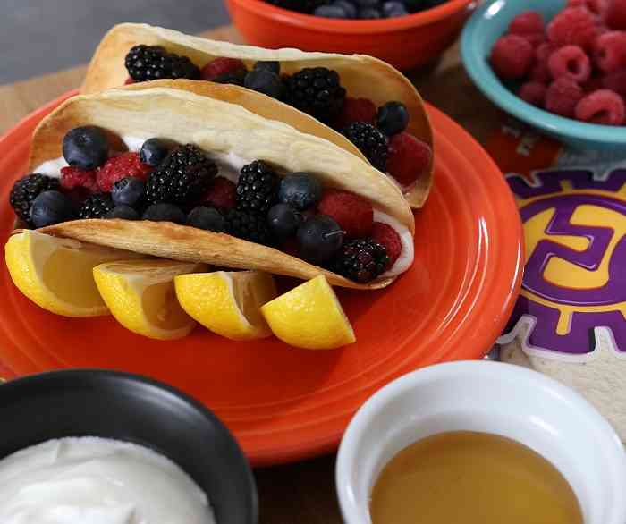Two fruit tacos made from Azteca Flour tortillas and fruit such as strawberries, blackberries, raspberries and blueberries on an orange plate held up by lemon wedges.