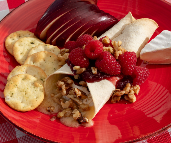 A baked brie on a red plate with apple slices, crackers, and some raspberries on top.